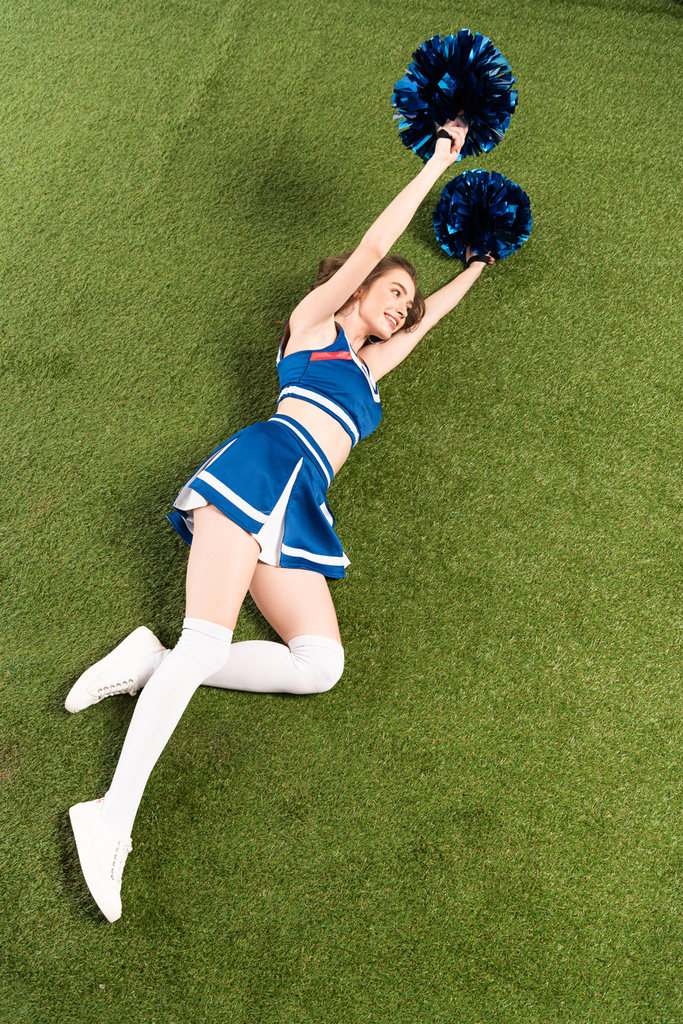 Widok z góry wesoły dziewczyna cheerleaderka w niebieskim mundurze leżącego z pomppoms na zielonym polu - Zdjęcie, obraz