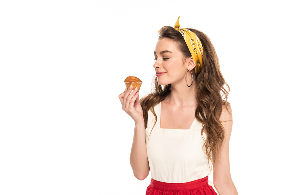 junge zufriedene Hausfrau in Kleid und Schürze mit Blick auf Muffin isoliert auf weiß - Foto, Bild