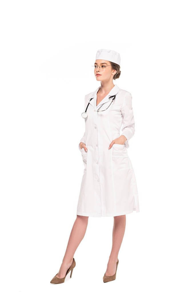 pohled plný délky mladého lékaře v bílém plášti se stetoskním pózem s rukama v kapsách izolovaných na bílém - Fotografie, Obrázek
