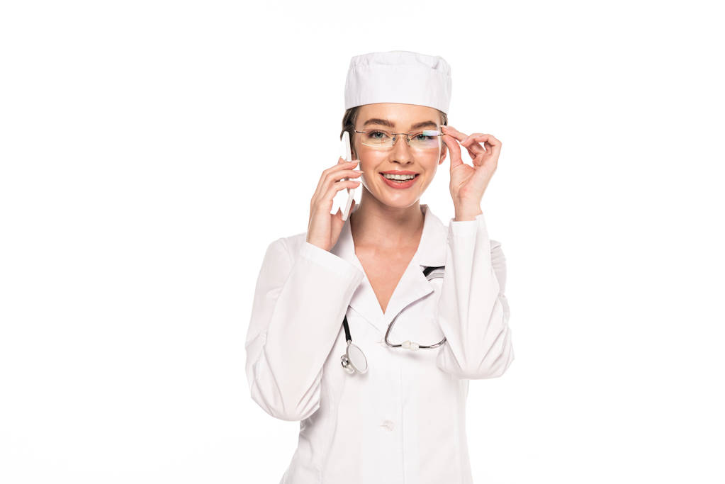 νέος χαμογελαστός γιατρός με λευκό παλτό μιλάει σε smartphone απομονωθεί σε λευκό - Φωτογραφία, εικόνα