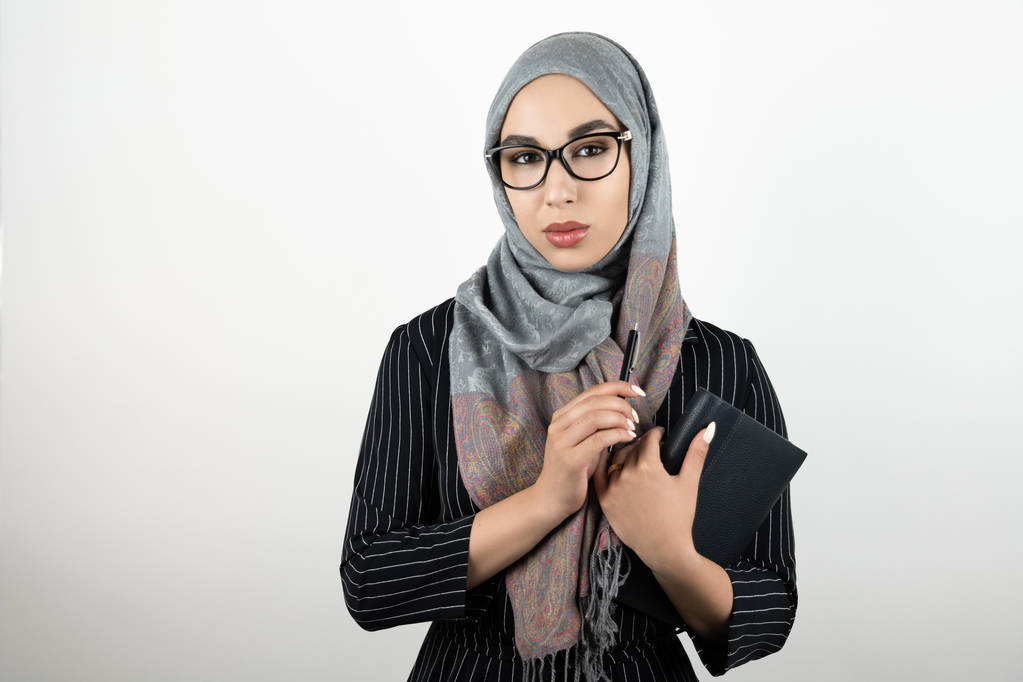 fiatal gyönyörű muszlim nő szemüveg viselése turbán hidzsáb, headsga kezében egy notebook és egy tollat elszigetelt fehér háttér - Fotó, kép