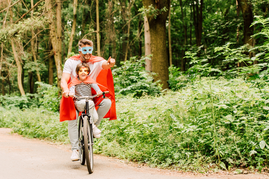 Vater und Kind fahren in Superheldenkostümen mit dem Fahrrad durch den Wald, während Papa mit dem Finger zeigt - Foto, Bild