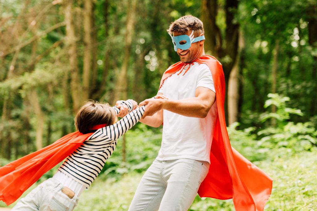 padre en capa y máscara girando niño pequeño en capa de superhéroe rojo
 - Foto, imagen