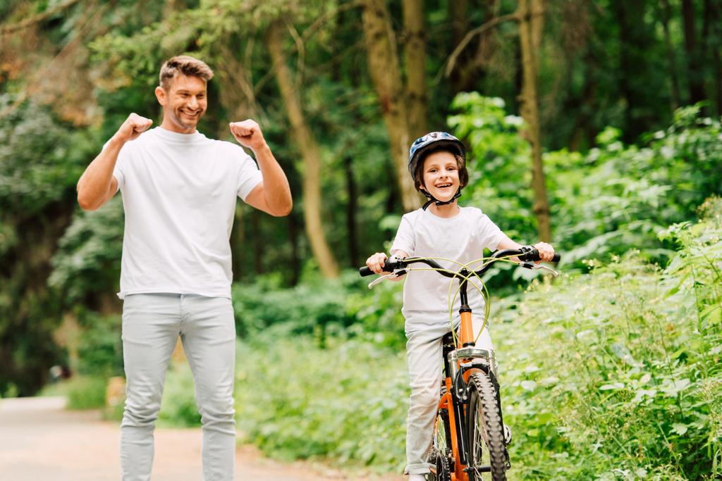 padre sonriendo y animando hijo mientras niño montar en bicicleta y mirando a la cámara
 - Foto, Imagen