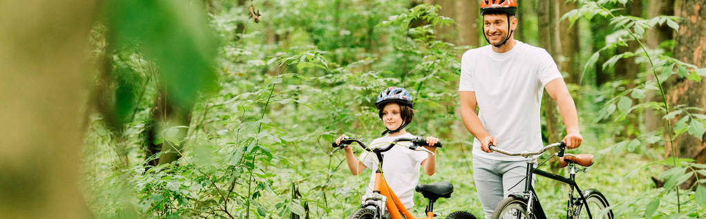 πανοραμική βολή του πατέρα και του γιου σε κράνη που περπατούν με ποδήλατα στο δάσος - Φωτογραφία, εικόνα