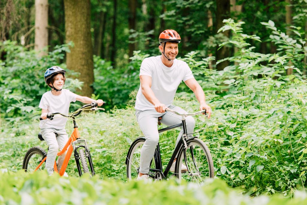 selectieve focus van vader en zoon glimlachend en Vooruitkijkend tijdens het rijden fietsen in bos - Foto, afbeelding
