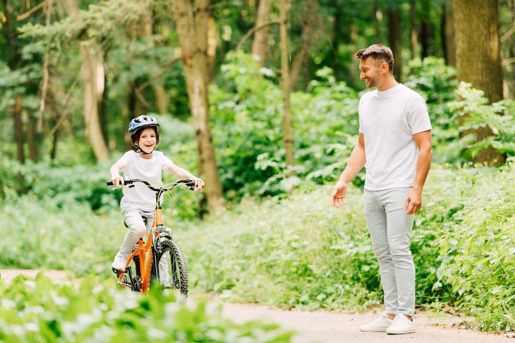Ganzkörperansicht des Sohnes, der Fahrrad fährt, und des Vaters, der neben dem Jungen steht und das Kind ansieht  - Foto, Bild