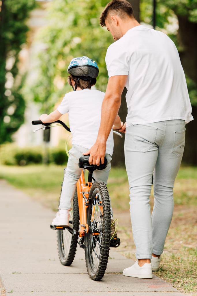 повний вигляд батька, який допомагає синові їздити на велосипеді, тримаючи сидіння на велосипеді
 - Фото, зображення