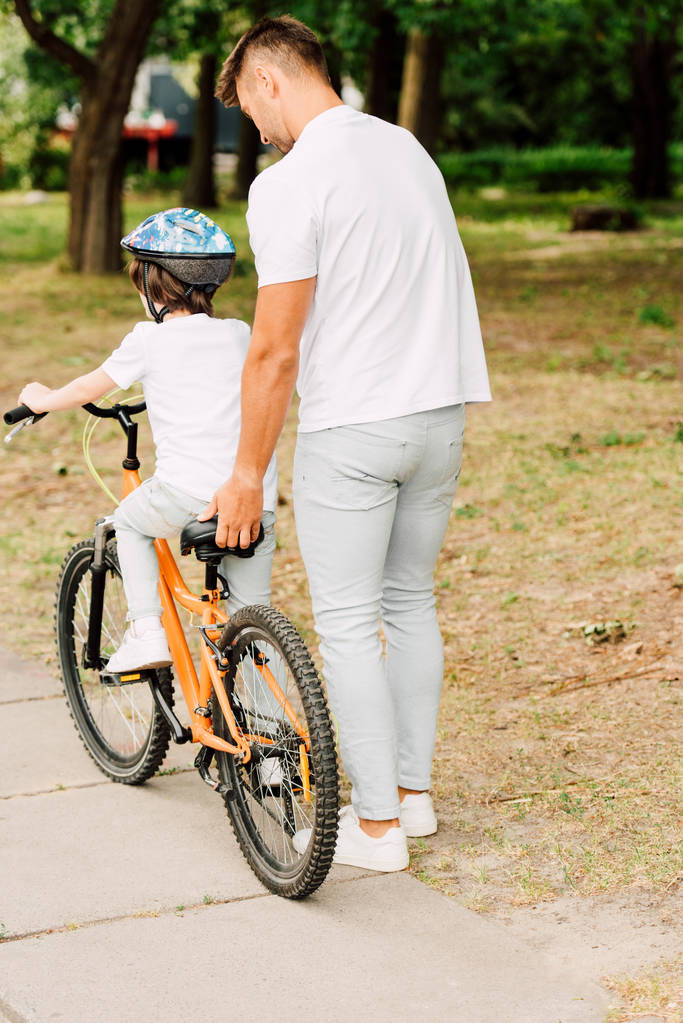 Rückansicht des Vaters, der seinem Sohn beim Fahrradfahren hilft, indem er den Sitz des Fahrrads hält - Foto, Bild