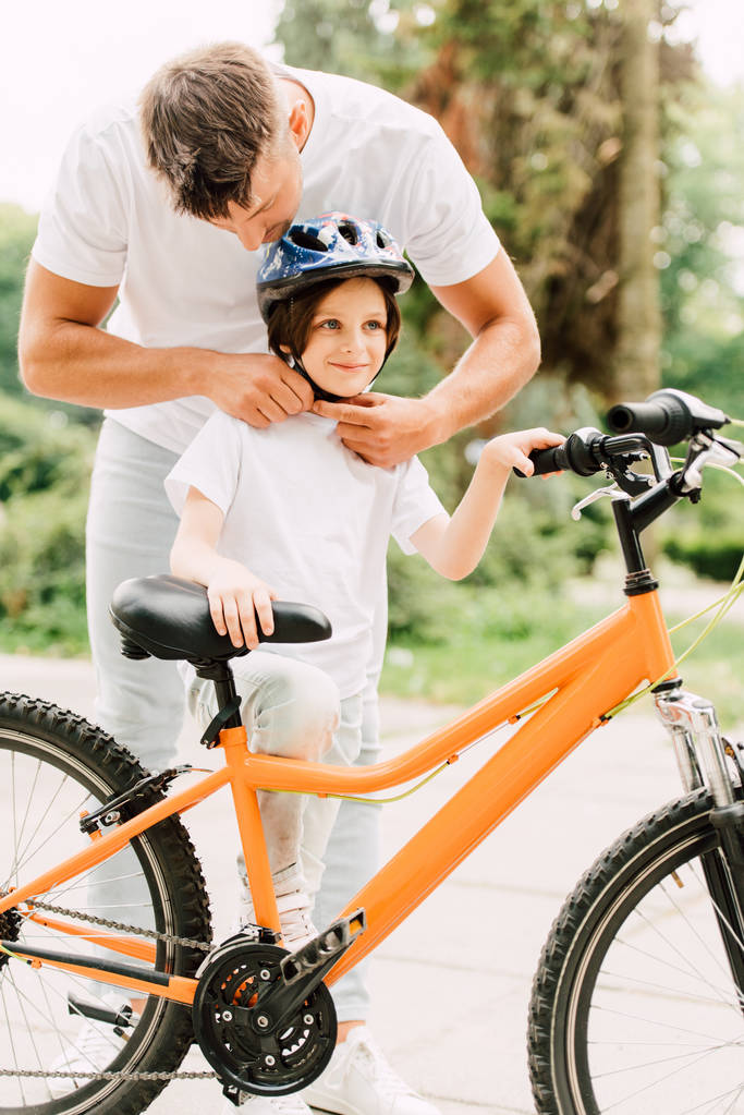 full length view of father putting helmet on son while boy standing near bicycle and looking away - Photo, Image