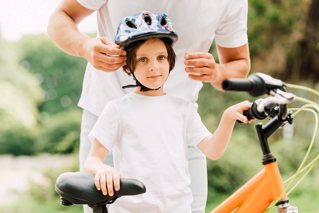 cropped view of boy looking at camera while father putting helmet on son - Photo, Image