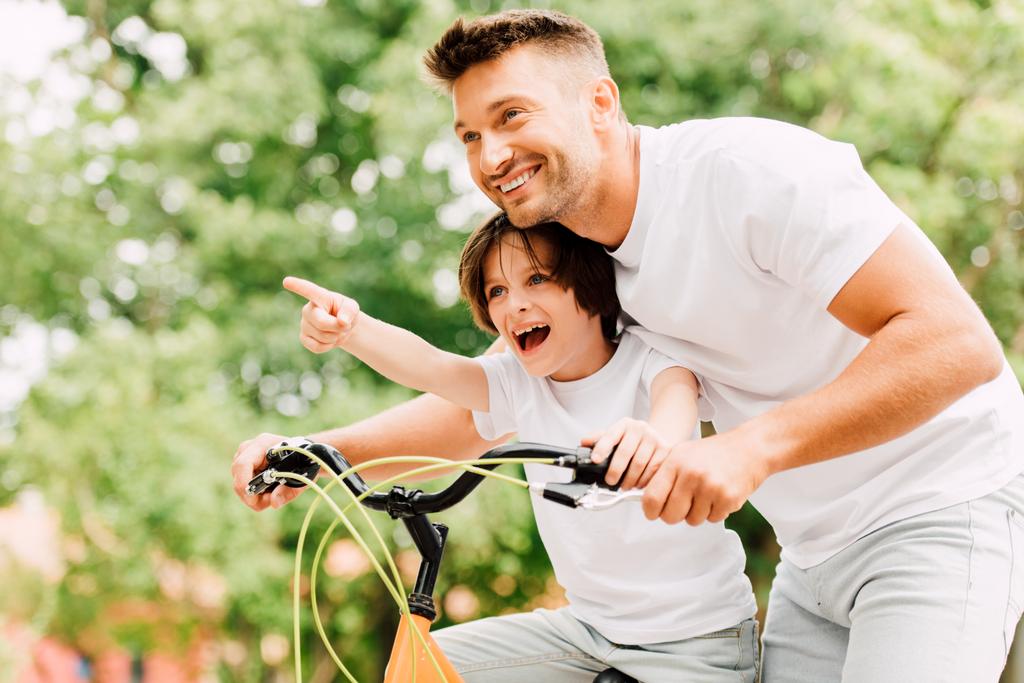 ενθουσιασμένος πατέρας και γιος κοιτάζοντας μπροστά, ενώ το αγόρι δείχνοντας με το δάχτυλο και τον μπαμπά βοηθώντας το παιδί να ιππεύσει στο ποδήλατο - Φωτογραφία, εικόνα