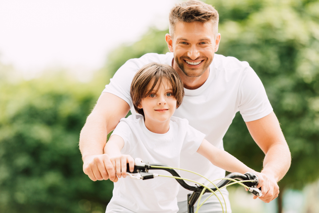Šťastný otec a syn se dívají na kameru zatímco chlapec se dívá na otce jízdního kola stojícího blízko dítěte - Fotografie, Obrázek