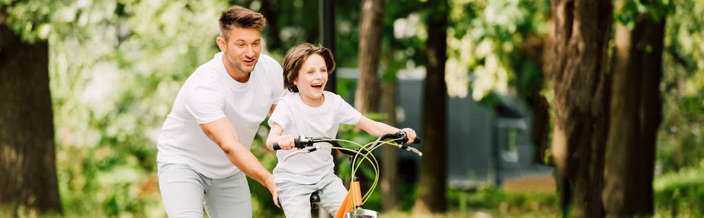 tiro panorámico de padre empujando bicicleta mientras el hijo montar en bicicleta
 - Foto, Imagen