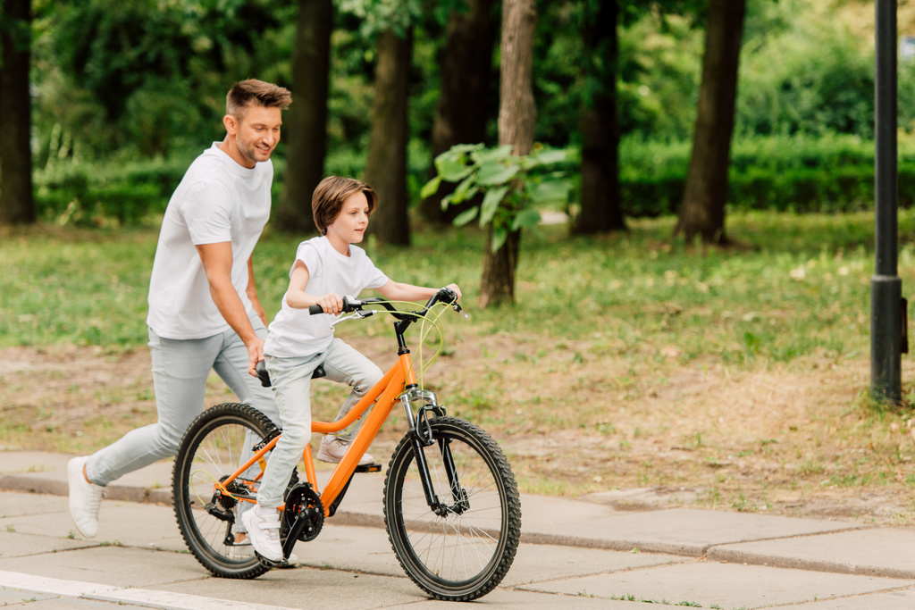 Вид отца в полный рост, толкающего велосипед и бегающего за сыном, пока ребенок катается на велосипеде
 - Фото, изображение