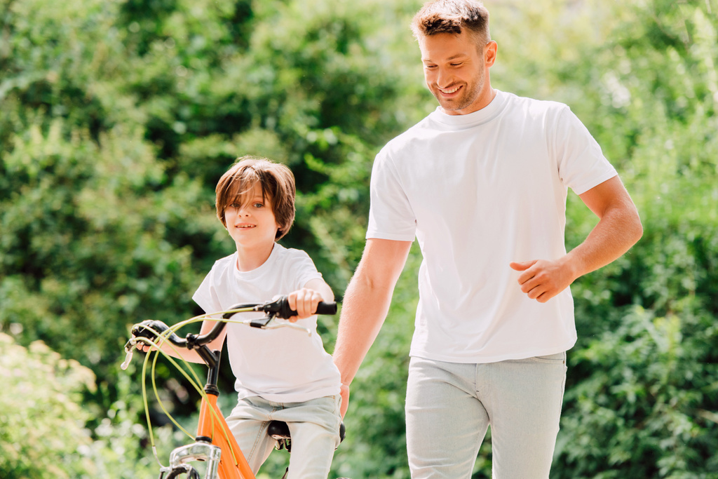 son riding bicycle and father walking next to kid and holding sit of bike  - Photo, Image