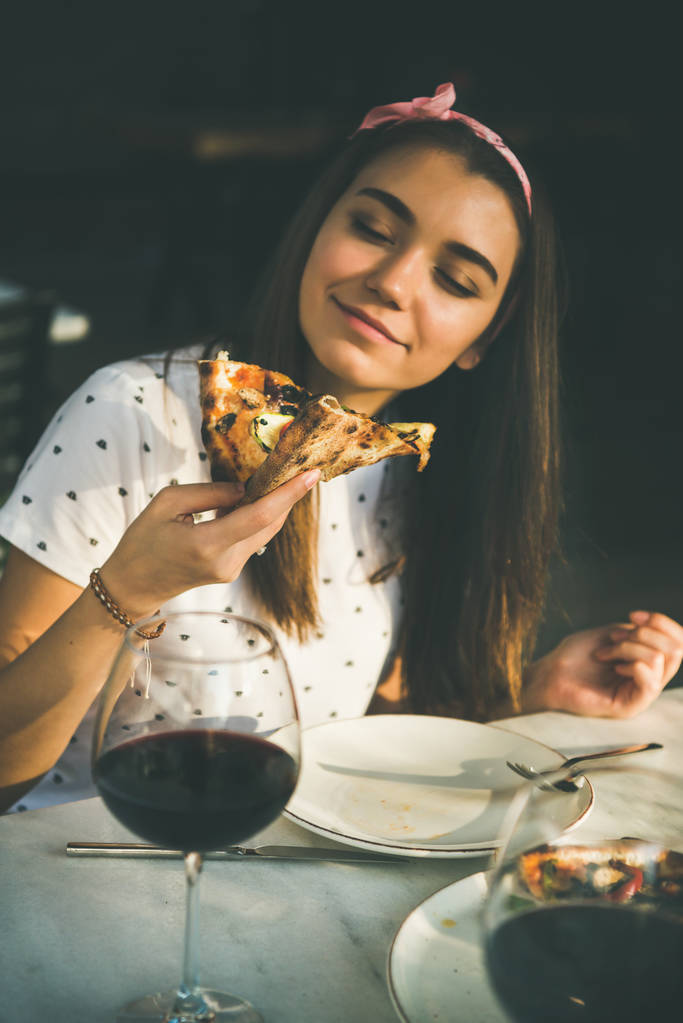 Joven mujer caucásica feliz sentado en la mesa comiendo pizza recién horneada y beber vino tinto en la cafetería de cocina italiana al aire libre en claro día soleado de verano
 - Foto, imagen