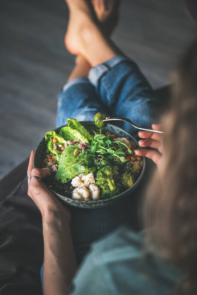 Zdravá večeře nebo oběd. Kudrnatá žena v tričku a džínách, které sedí doma a jedí Superbowlu veganové nebo Buddha mísu s hummus, zeleninou, čerstvým salátem, fazolemi, kuskly a avokádem, výhled - Fotografie, Obrázek