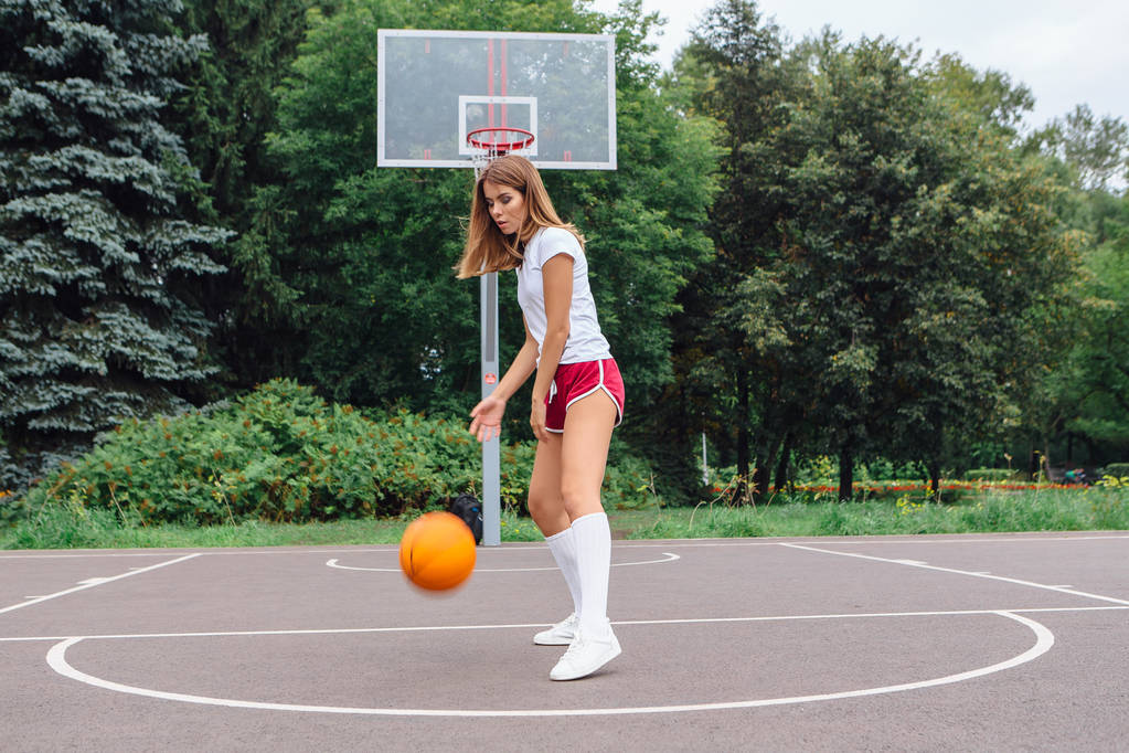Красивая молодая девушка в белой футболке, шортах и кроссовках, играет с мячом на баскетбольной площадке
. - Фото, изображение