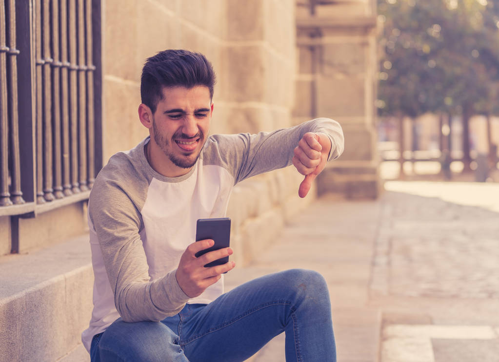Portrait of young student man reacting negative to post in social media network or dating app with dislike showing thumb down. In social network rating likes, reactions and reassurance youth problems. - Photo, Image