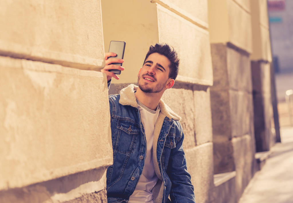 Emocionado joven de moda tomando una selfie en el teléfono móvil inteligente en vacaciones en la calle de la ciudad europea o en el campus universitario fuera. En vacaciones y tecnología, conexión y concepto de redes sociales
. - Foto, Imagen