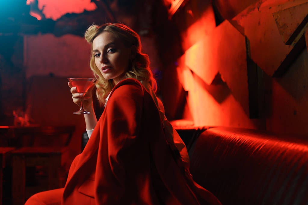 Wizerunek młodej Blondynki w czerwonej kurtce patrząc na kamerę z koktajlem w ręku w klubie nocnym - Zdjęcie, obraz