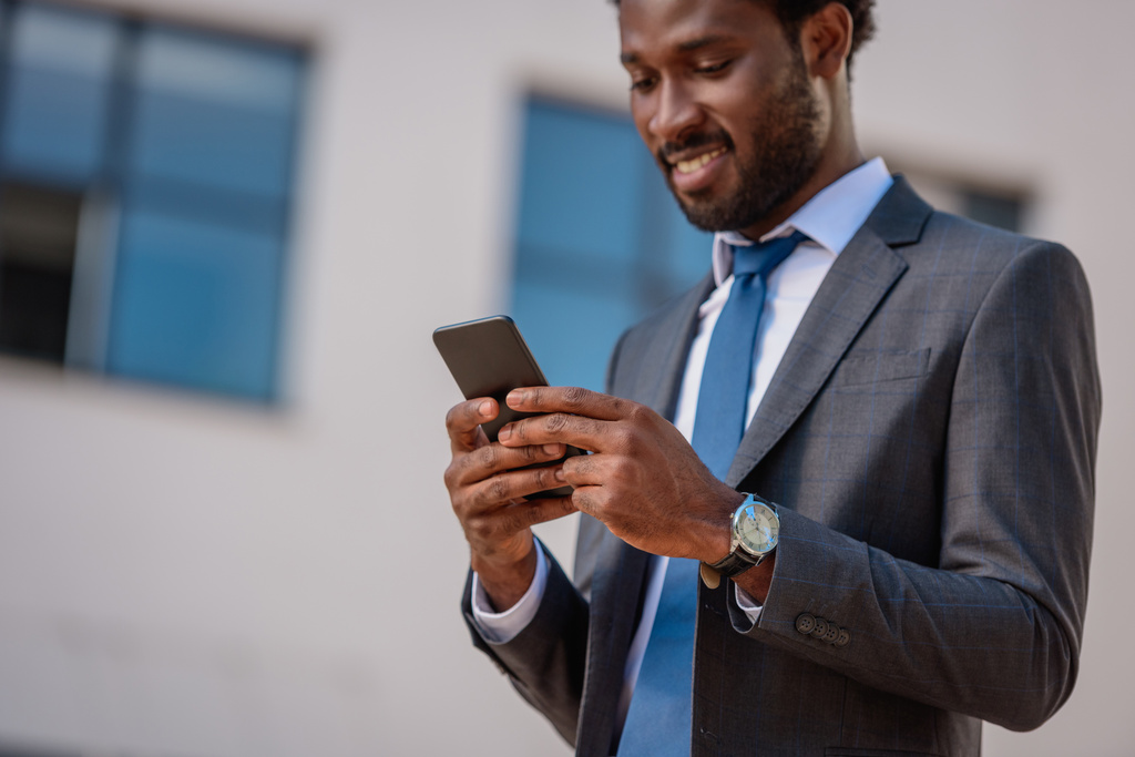 スマートフォンを使用しながら笑顔の陽気なアフリカ系アメリカ人ビジネスマン - 写真・画像