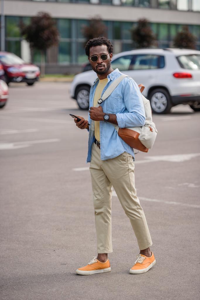 bell'uomo afro-americano in occhiali da sole che tiene lo smartphone mentre si trova sul parcheggio auto
 - Foto, immagini