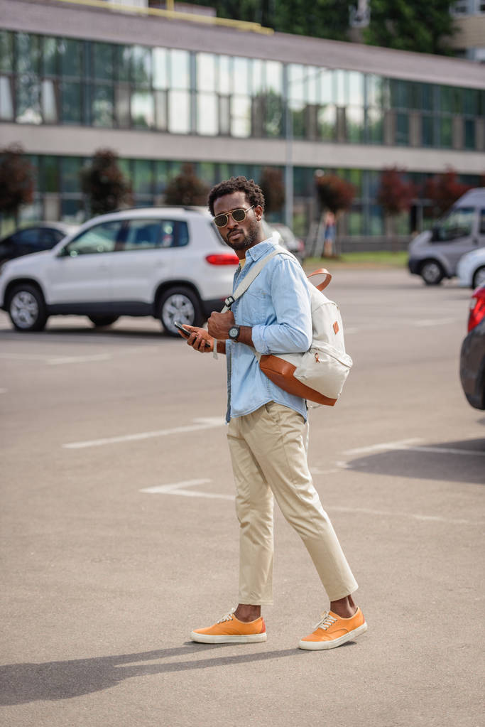 駐車場に立ってスマートフォンを持っている間、カメラを見ているハンサムなアフリカ系アメリカ人の男 - 写真・画像