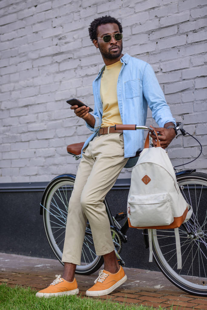 Stylový Afroameričan, který drží telefon a dívá se jinam, zatímco stojí u jízdního kola poblíž zdi - Fotografie, Obrázek
