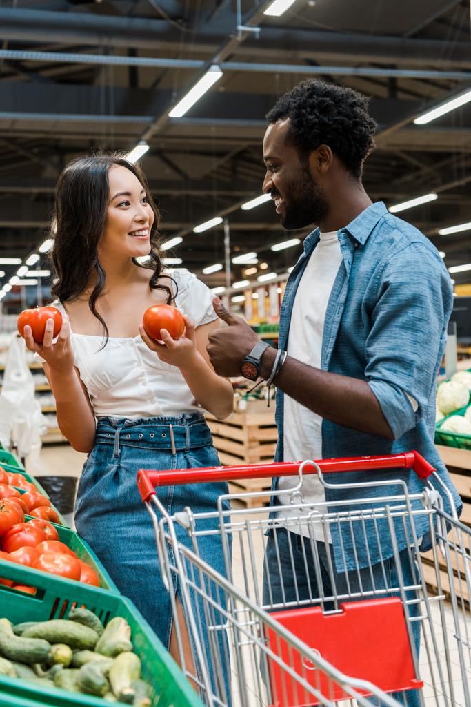 szelektív középpontjában ázsiai nő gazdaság friss paradicsom közelében vidám afro-amerikai ember mutatja hüvelykujját fel szupermarket  - Fotó, kép