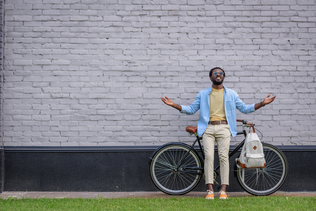 vzrušený afroamerický muž gestikuluje, zatímco stojí u cihlové zdi s bicyklem - Fotografie, Obrázek