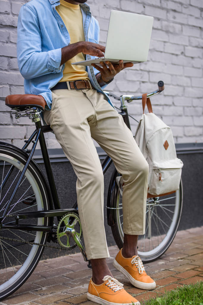 μερική θέα του αφρικανικού αμερικανικού ανθρώπου χρησιμοποιώντας φορητό υπολογιστή ενώ στέκεται κοντά στο ποδήλατο - Φωτογραφία, εικόνα