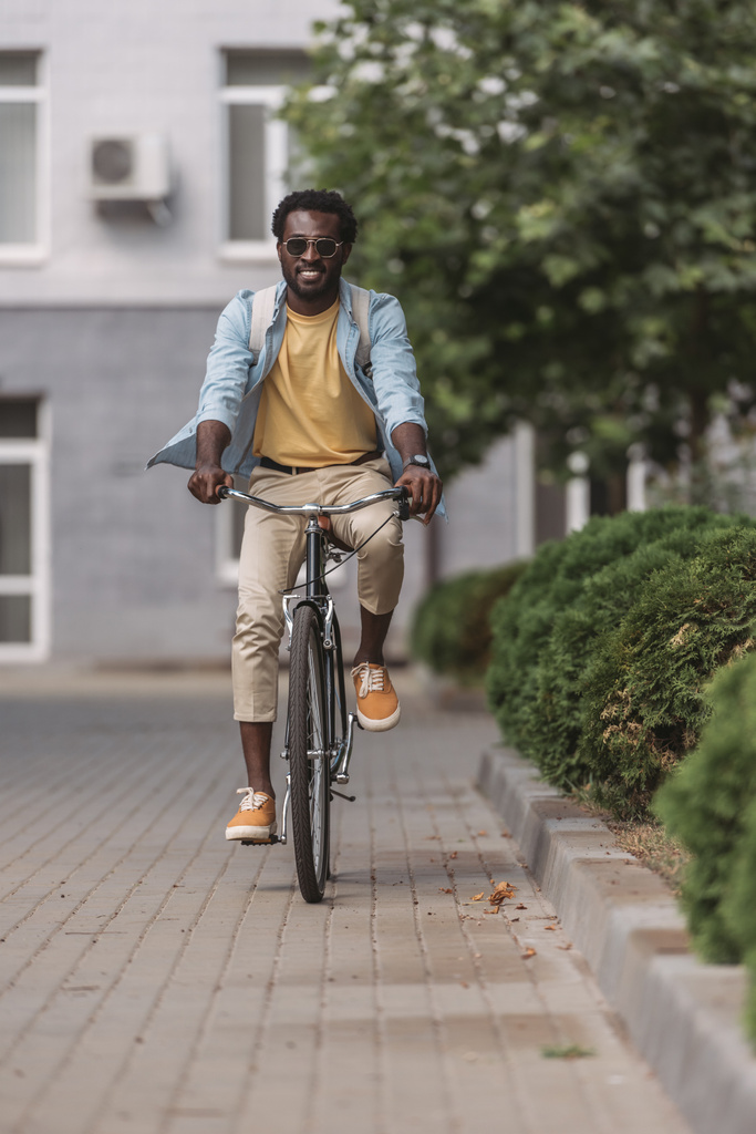 красивий, стильний афроамериканський чоловік посміхається під час їзди на велосипеді вздовж вулиці
 - Фото, зображення