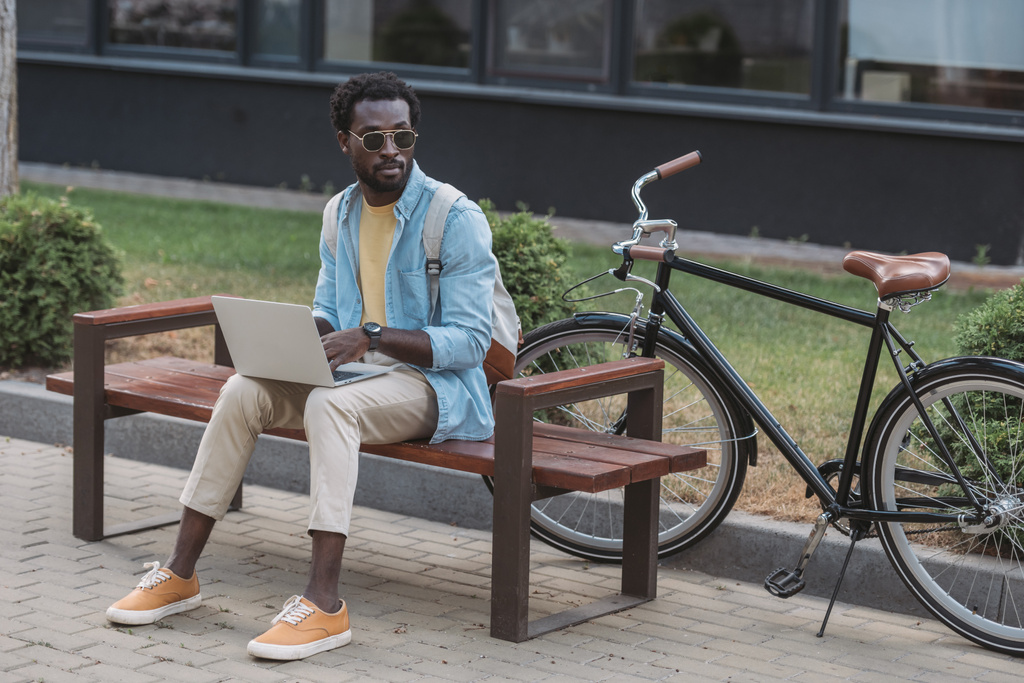 schöner afrikanisch-amerikanischer Mann mit Laptop, während er auf einer Bank in der Nähe von Fahrrad sitzt und wegschaut - Foto, Bild