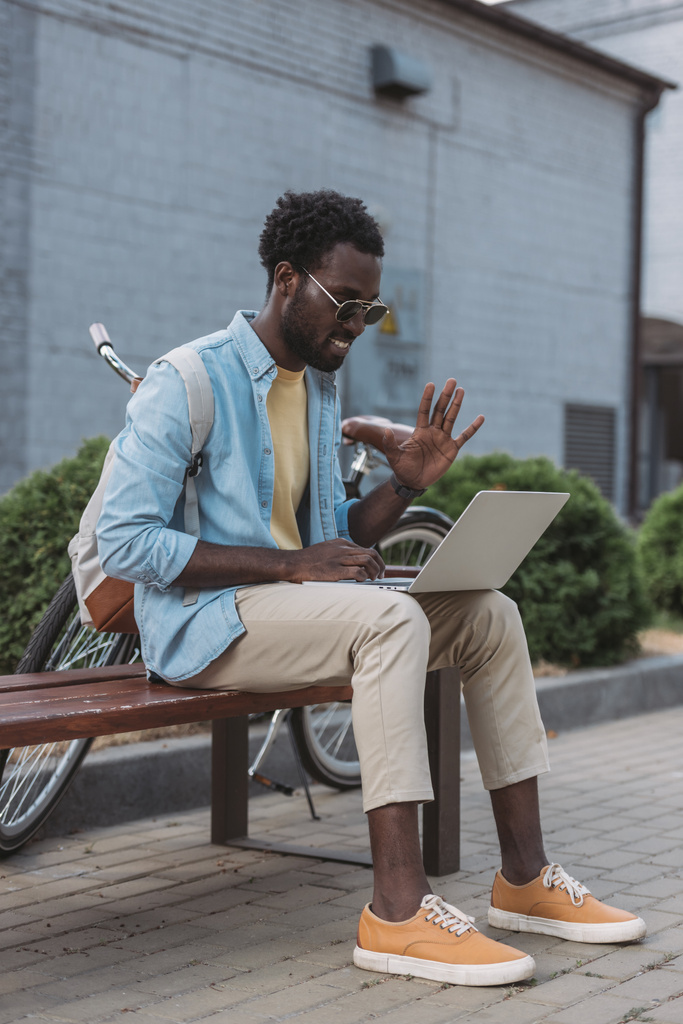 веселий афроамериканський чоловік махає рукою, сидячи на лавці під час відеодзвінка в ноутбуці
 - Фото, зображення
