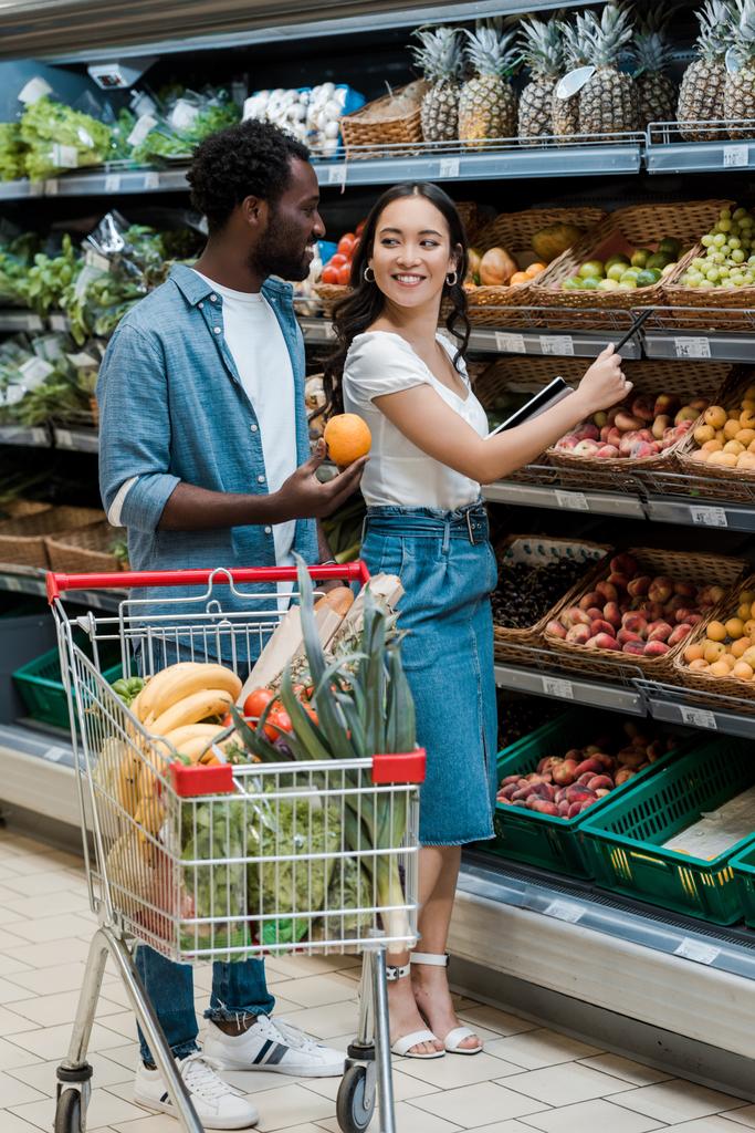 szczęśliwy African amerykański mężczyzna patrząc na Asian kobieta stojący w supermarket blisko owoce  - Zdjęcie, obraz