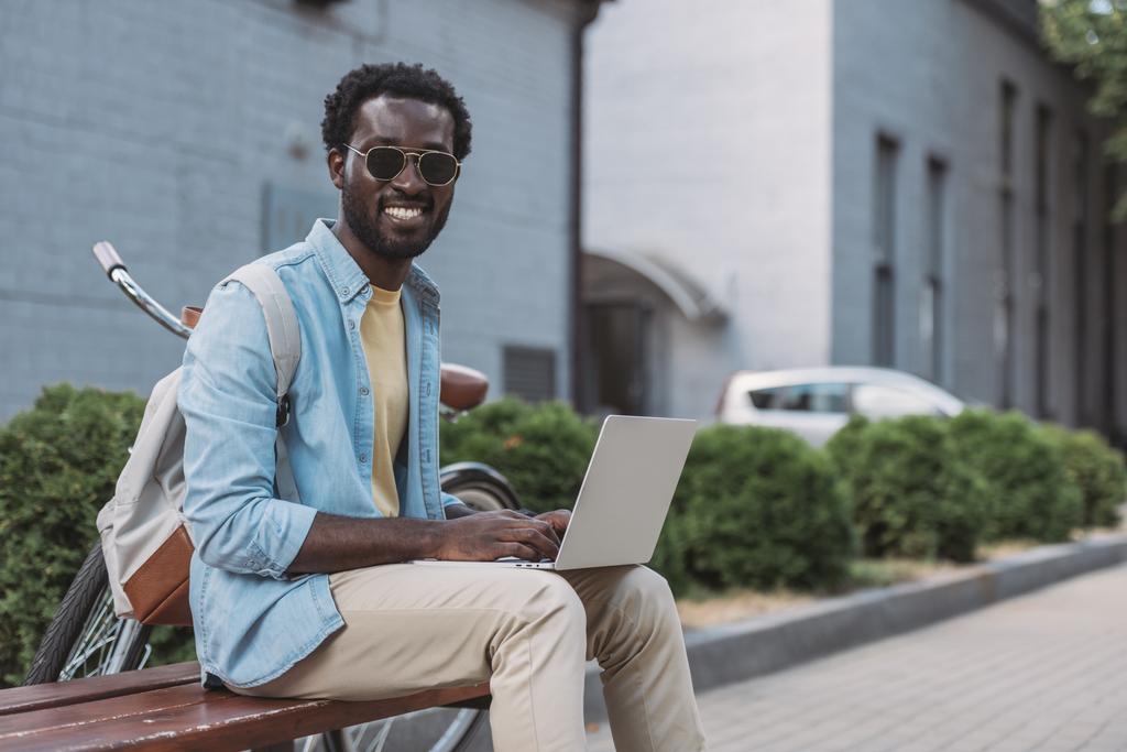 iloinen afrikkalainen amerikkalainen mies hymyilee kameralle istuessaan penkillä ja käyttämällä kannettavaa tietokonetta
 - Valokuva, kuva