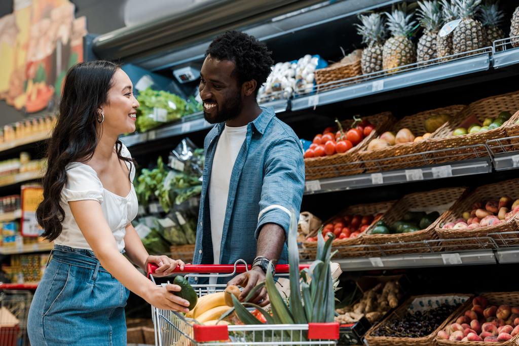 mutlu asyalı kız mağazada alışveriş sepeti ve Afro-Amerikan adam yakınında avokado tutan  - Fotoğraf, Görsel