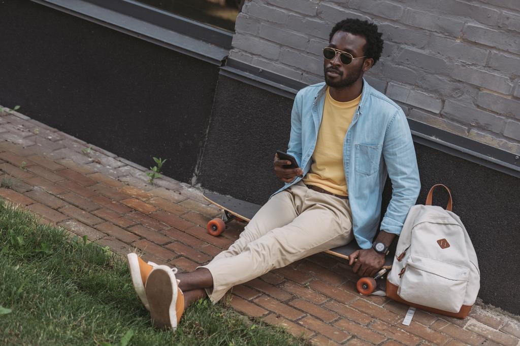 csinos, stílusos afro-amerikai ember ül Longboard és a gazdaság okostelefon - Fotó, kép