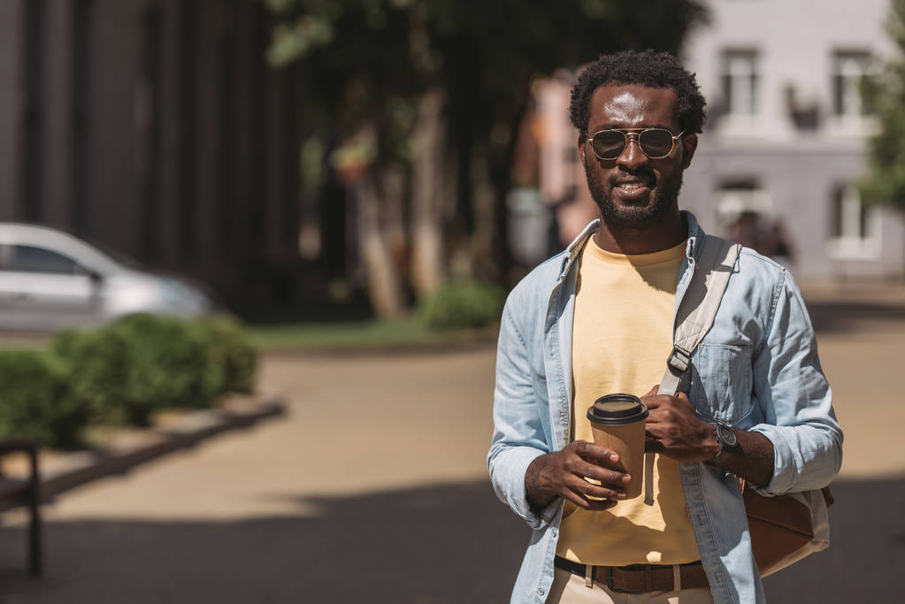pohledný, stylový afroamerický muž v brýlích přidržel papírový pohár při pohledu na fotoaparát - Fotografie, Obrázek