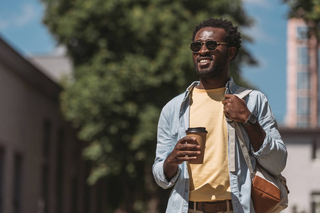θετικός, κομψός Αφρικανός Αμερικανός που κρατά καφέ για να πάει και να κοιτάει μακριά - Φωτογραφία, εικόνα