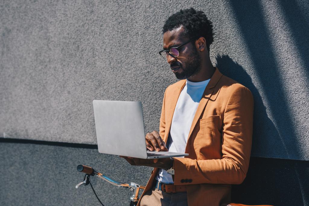 uważny African American biznesmen w okularach przy użyciu laptopa stojąc na ścianie z rowerem - Zdjęcie, obraz
