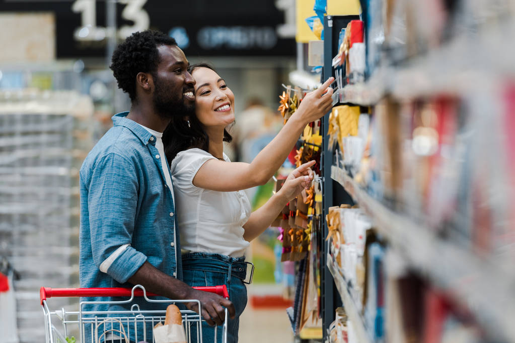 selectieve focus van gelukkige Afro-Amerikaanse man staand met Aziatische vrouw die met de vinger naar boodschappen in de supermarkt wijst  - Foto, afbeelding