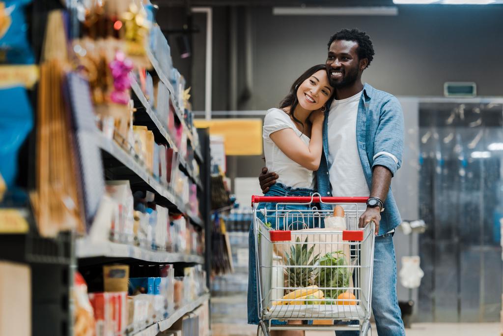 enfoque selectivo de alegre africano americano hombre abrazando asiático novia en supermercado
  - Foto, Imagen
