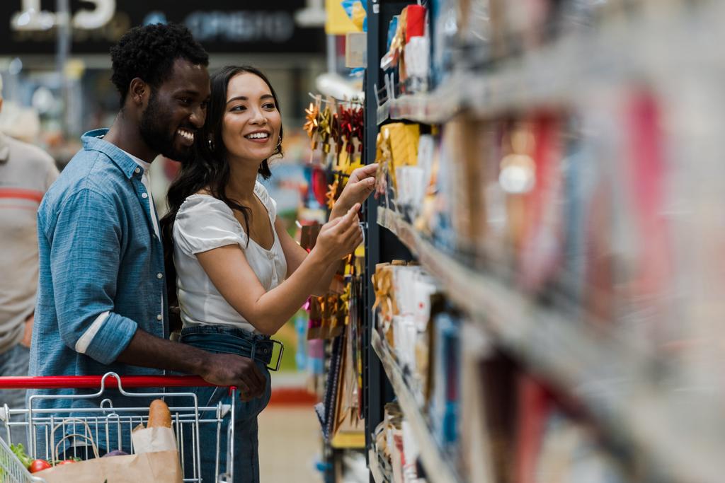 スーパーマーケットで食料品を指差すアジアの女性と立っている陽気なアフリカ系アメリカ人男性の選択的な焦点  - 写真・画像
