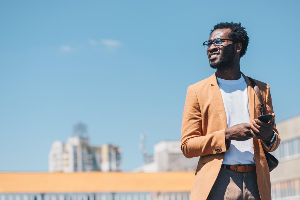 スマートフォンを使用して陽気なアフリカ系アメリカ人のビジネスマンと背景に青い空で微笑む - 写真・画像