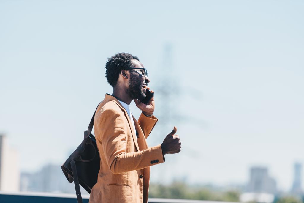 陽気なアフリカ系アメリカ人のビジネスマンがスマートフォンで話し、屋上を歩きながら親指を上げる  - 写真・画像