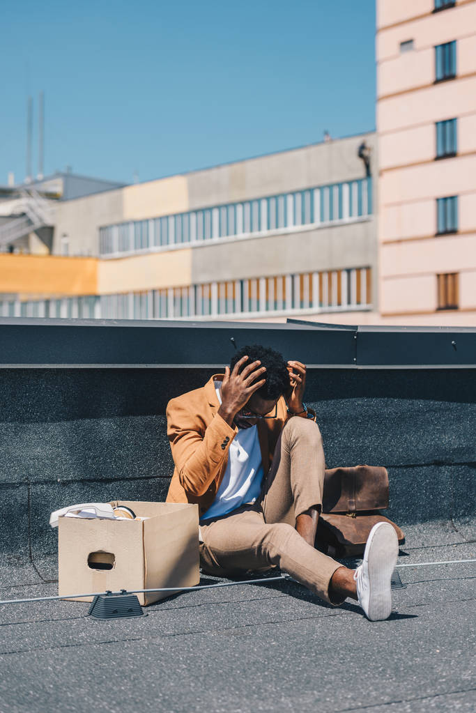 落ち込んだアフリカ系アメリカ人のビジネスマンは、カートンボックスの近くの屋上に座って、頭の近くに手をつないでいる - 写真・画像