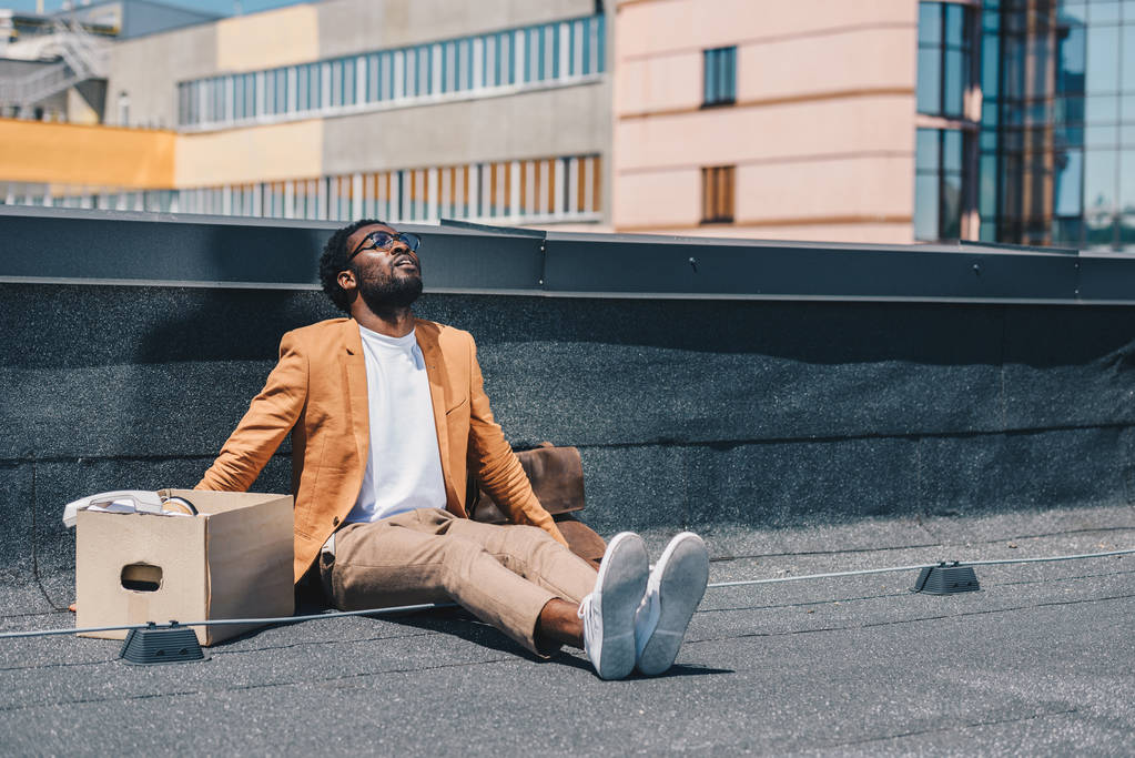 hombre de negocios afroamericano deprimido y despedido mirando hacia arriba mientras estaba sentado en la azotea cerca de la caja de cartón
 - Foto, Imagen